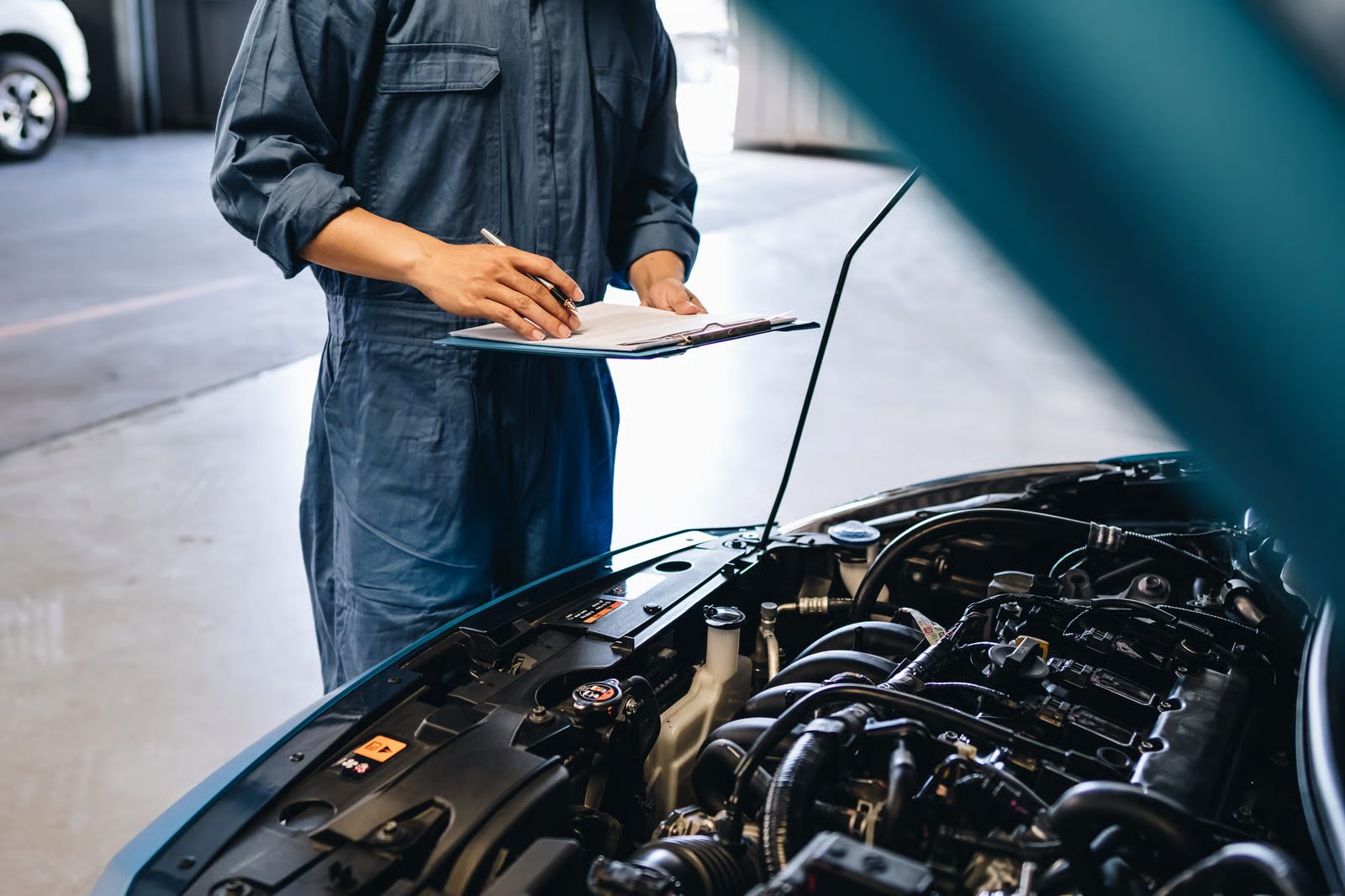 Auto mechanic in blue coveralls holding a clipboard and checklist while inspecting an open car engine bay in a repair shop