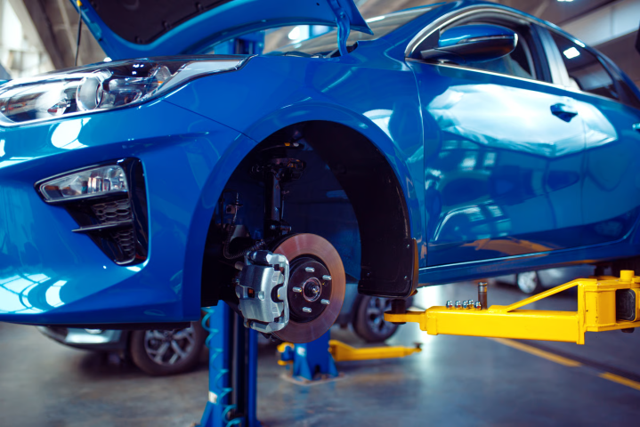Blue car elevated on a workshop lift with front wheel removed showing disc brake assembly and suspension components