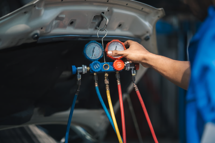Technician adjusting an air conditioning manifold gauge set hanging from the open car hood during an AC system pressure test